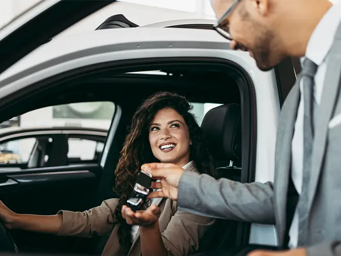 photo of a car salesman handing car keys to a smiling woman sitting in the driver's seat of a car