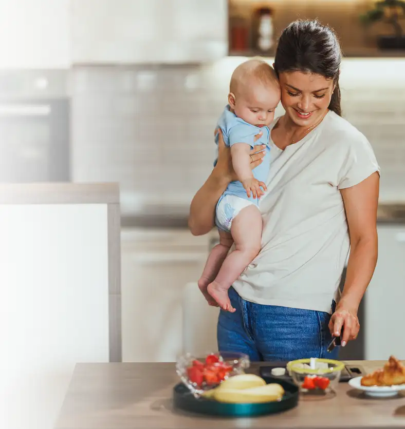 mom-cooking-with-baby-iStock-2214374097 photo of a mom holding her baby while getting food ready in the kitchen